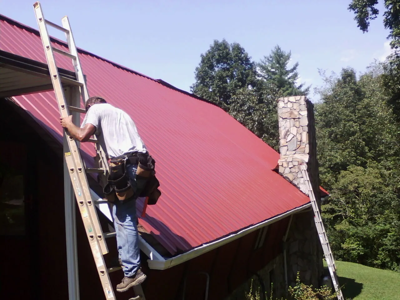 Roofer on a ladder beside a red metal roof and stone chimney — Soffit & Fascia Repair by Roofing Brothers in Kinderhook